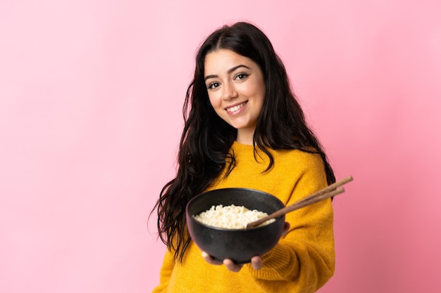 Jovem mulher caucasiana isolada em um fundo rosa com uma expressão feliz, segurando uma tigela de macarrão com pauzinhos