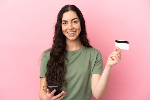 Jovem mulher caucasiana isolada em um fundo rosa comprando com o celular com um cartão de crédito
