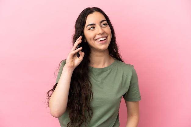 Jovem mulher caucasiana isolada em um fundo rosa, conversando com alguém ao telefone celular