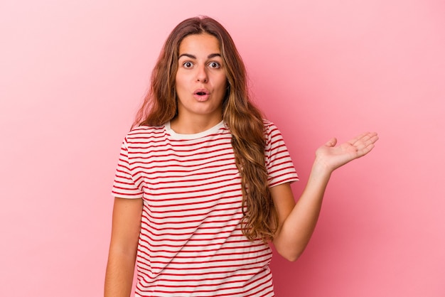 Jovem mulher caucasiana isolada em um fundo rosa impressionado segurando o espaço da cópia na palma da mão.