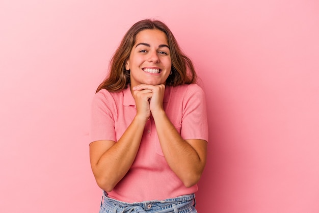 Jovem mulher caucasiana isolada em um fundo rosa mantém as mãos sob o queixo, olhando alegremente para o lado.