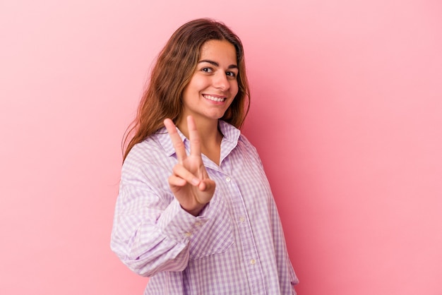 Jovem mulher caucasiana isolada em um fundo rosa, mostrando sinal de vitória e sorrindo amplamente.