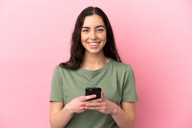 Jovem mulher caucasiana isolada em um fundo rosa olhando para a câmera e sorrindo enquanto usa o celular