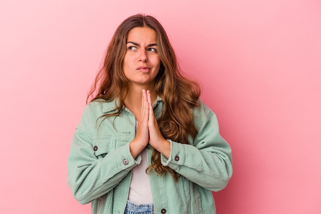 Jovem mulher caucasiana isolada em um fundo rosa orando, mostrando devoção, pessoa religiosa em busca de inspiração divina.