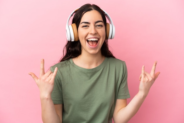 Jovem mulher caucasiana isolada em um fundo rosa ouvindo música fazendo gesto de rock