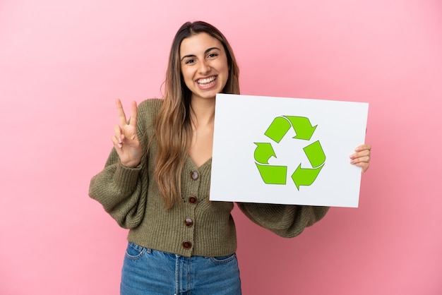 Jovem mulher caucasiana isolada em um fundo rosa segurando um cartaz com o ícone de reciclagem e comemorando uma vitória