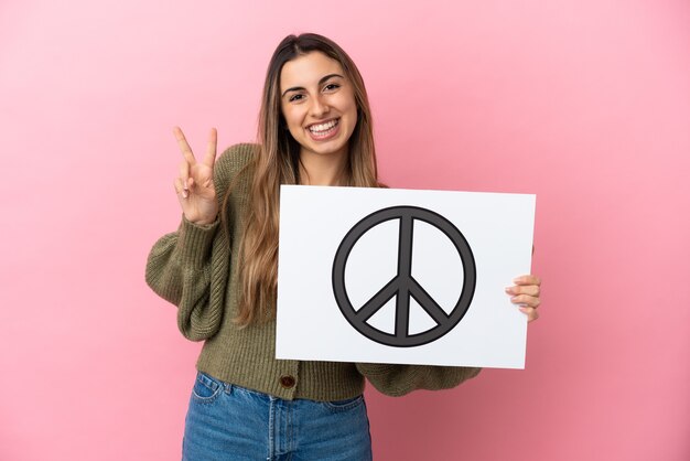 Jovem mulher caucasiana isolada em um fundo rosa segurando um cartaz com o símbolo da paz e comemorando uma vitória