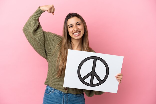 Jovem mulher caucasiana isolada em um fundo rosa segurando um cartaz com o símbolo da paz e fazendo um gesto forte