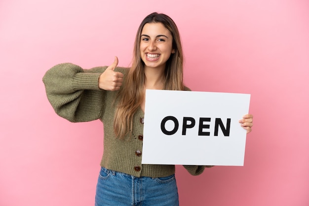 Jovem mulher caucasiana isolada em um fundo rosa segurando um cartaz com o texto ABRIR com o polegar para cima