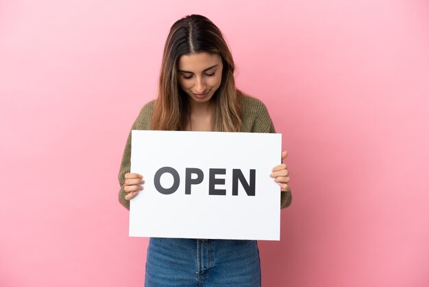 Jovem mulher caucasiana isolada em um fundo rosa segurando um cartaz com o texto ABRIR