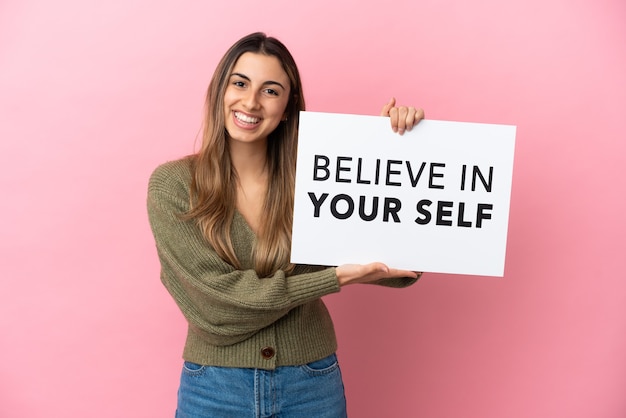 Jovem mulher caucasiana isolada em um fundo rosa segurando um cartaz com o texto Acredite em si mesmo e apontando-o