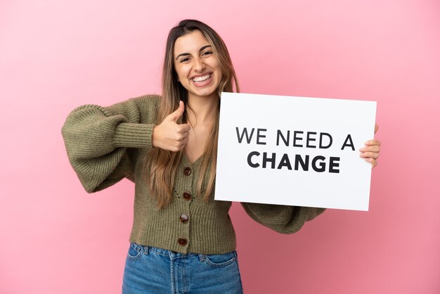 Jovem mulher caucasiana isolada em um fundo rosa segurando um cartaz com o texto Precisamos de uma mudança com o polegar para cima