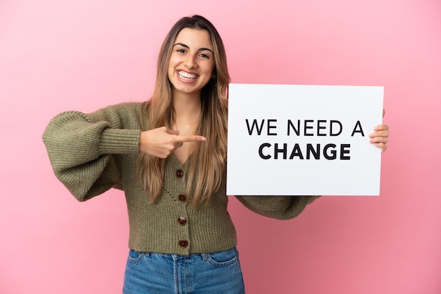 Jovem mulher caucasiana isolada em um fundo rosa segurando um cartaz com o texto Precisamos de uma mudança e apontando-o