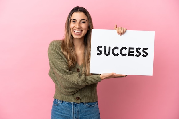 Jovem mulher caucasiana isolada em um fundo rosa segurando um cartaz com o texto SUCESSO com uma expressão feliz