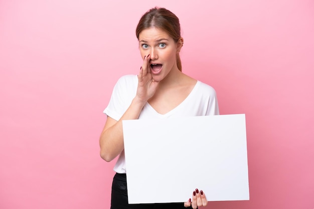 Jovem mulher caucasiana isolada em um fundo rosa segurando um cartaz vazio e gritando