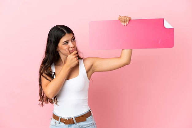 Jovem mulher caucasiana isolada em um fundo rosa segurando um cartaz vazio e pensando