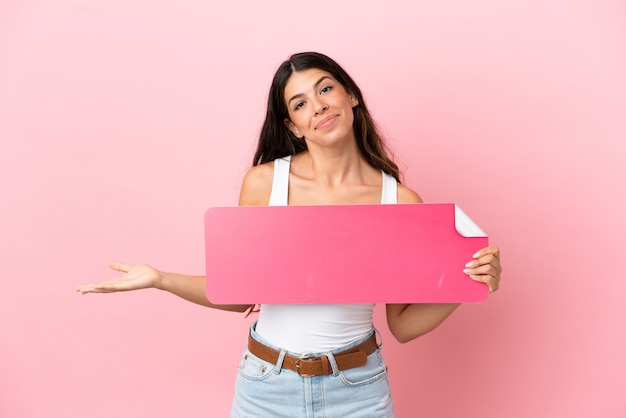 Jovem mulher caucasiana isolada em um fundo rosa segurando um cartaz vazio e tendo dúvidas