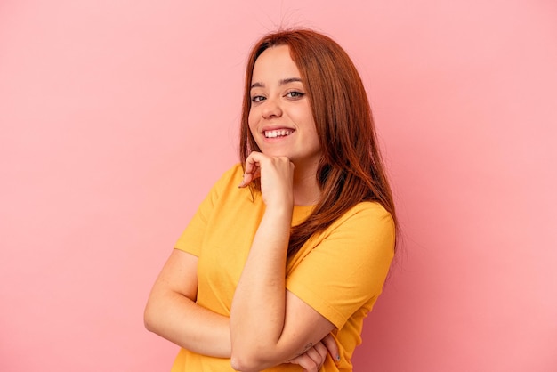 Jovem mulher caucasiana isolada em um fundo rosa, sorrindo feliz e confiante, tocando o queixo com a mão.