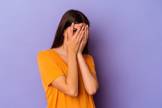 Jovem mulher caucasiana isolada em um fundo roxo piscar para a câmera por entre os dedos, o rosto coberto de vergonha.