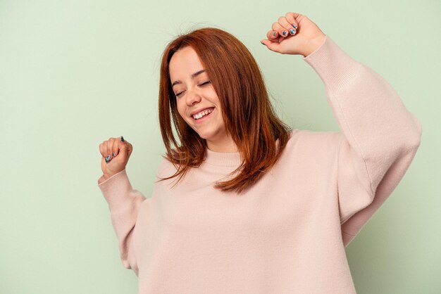 Jovem mulher caucasiana isolada em um fundo verde, comemorando um dia especial, pula e levanta os braços com energia.