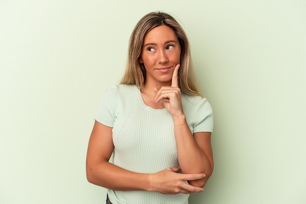 Jovem mulher caucasiana isolada em um fundo verde, contemplando, planejando uma estratégia, pensando na forma de um negócio.