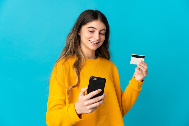 Jovem mulher caucasiana isolada em uma parede azul comprando com o celular com um cartão de crédito