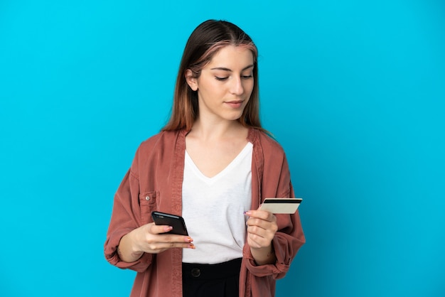 Jovem mulher caucasiana isolada em uma parede azul comprando com o celular com um cartão de crédito