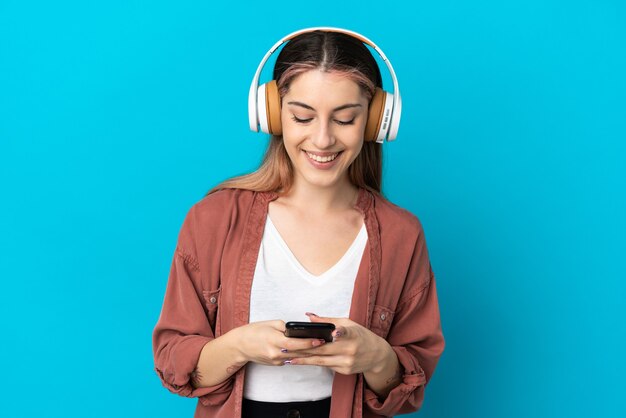 Jovem mulher caucasiana isolada em uma parede azul ouvindo música e olhando para o celular