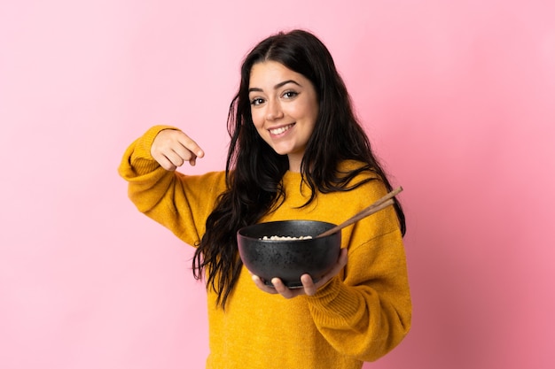 Jovem mulher caucasiana isolada em uma parede rosa e apontando enquanto segura uma tigela de macarrão com pauzinhos