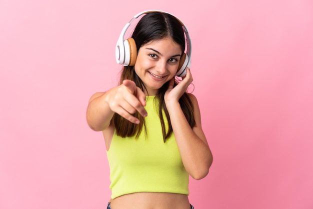 Jovem mulher caucasiana isolada em uma parede rosa ouvindo música e apontando para a frente