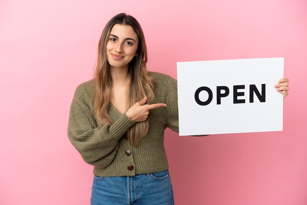 Jovem mulher caucasiana isolada em uma parede rosa segurando um cartaz com o texto ABRIR e apontando-o