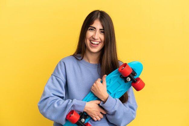 Jovem mulher caucasiana isolada em uma superfície amarela com um skate e uma expressão feliz