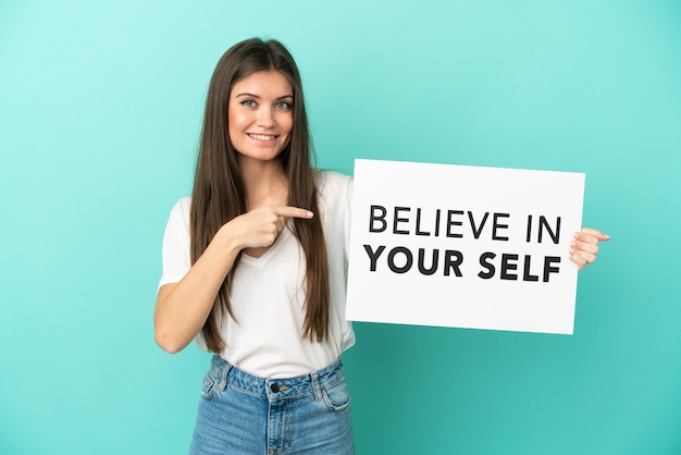 Jovem mulher caucasiana isolada na parede azul segurando um cartaz com o texto Acredite em si mesmo e apontando-o