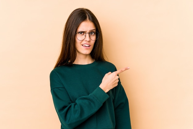 Jovem mulher caucasiana isolada na parede bege, sorrindo e apontando de lado, mostrando algo no espaço em branco.