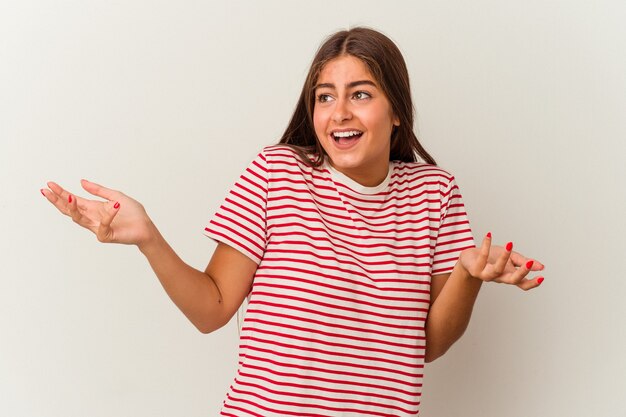 Jovem mulher caucasiana, isolada na parede branca, rezando para ter sorte, espantada e abrindo a boca, olhando para a frente.