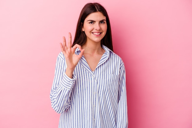 Jovem mulher caucasiana isolada na parede rosa pisca um olho e segura um gesto de ok com a mão.