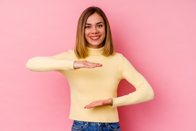 Jovem mulher caucasiana isolada na parede rosa segurando algo com as duas mãos, apresentação do produto.