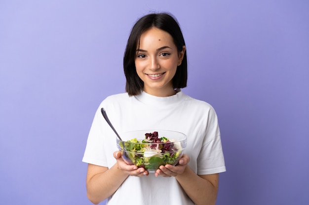 Jovem mulher caucasiana isolada na parede roxa segurando uma tigela de salada com uma expressão feliz