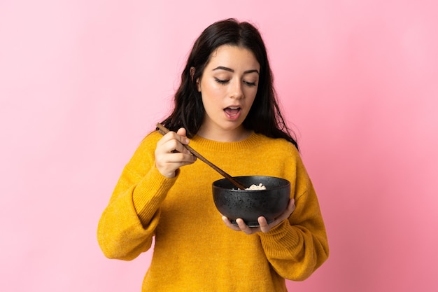 Jovem mulher caucasiana isolada na rosa segurando uma tigela de macarrão com pauzinhos