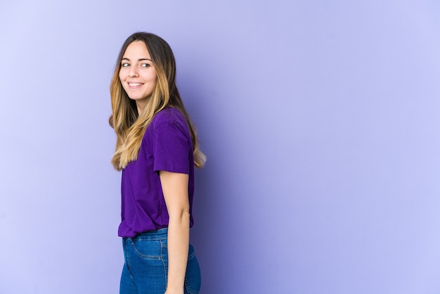 Jovem mulher caucasiana isolada no espaço roxo parece de lado sorrindo, alegre e agradável.