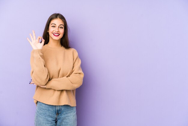 Jovem mulher caucasiana isolada no espaço roxo pisca um olho e segura um gesto de ok com a mão.