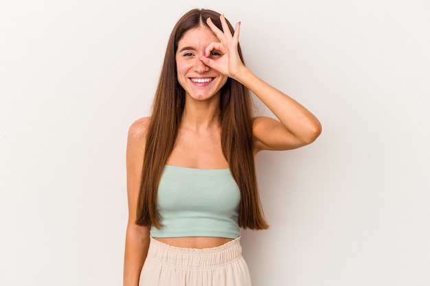 Jovem mulher caucasiana isolada no fundo branco animada mantendo o gesto ok no olho.