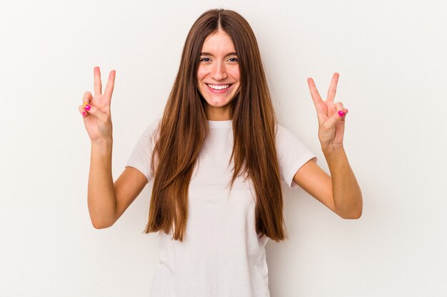 Jovem mulher caucasiana isolada no fundo branco, mostrando o sinal da vitória e sorrindo amplamente.