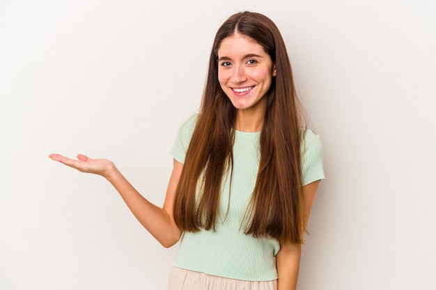 Jovem mulher caucasiana isolada no fundo branco, mostrando um espaço de cópia na palma da mão e segurando a outra mão na cintura.