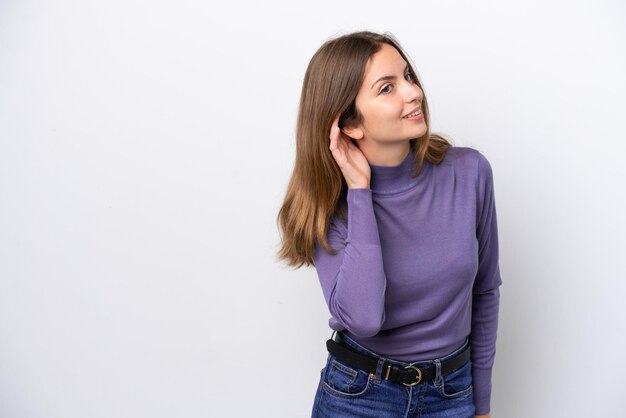 Jovem mulher caucasiana isolada no fundo branco ouvindo algo colocando a mão na orelha