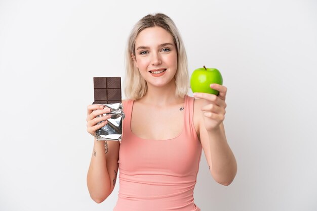 Jovem mulher caucasiana isolada no fundo branco pegando um tablete de chocolate em uma mão e uma maçã na outra