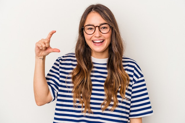 Jovem mulher caucasiana, isolada no fundo branco, segurando algo pequeno com os indicadores, sorrindo e confiante.