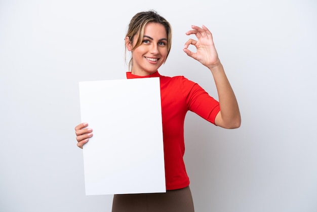 Jovem mulher caucasiana isolada no fundo branco segurando um cartaz vazio e fazendo sinal de OK