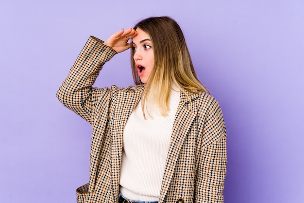 Jovem mulher caucasiana isolada no fundo roxo, olhando para longe, mantendo a mão na testa.