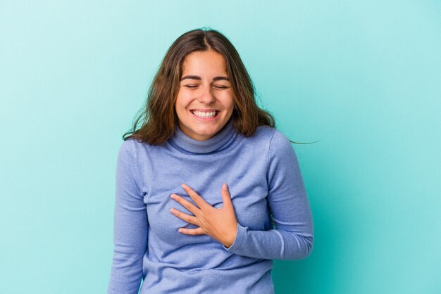 Jovem mulher caucasiana isolada sobre fundo azul ri alto, mantendo a mão no peito.